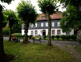 Historisches Gebäude mit Schieferfassade und rotem Ziegeldach, umgeben von Bäumen und Bänken im Vordergrund.