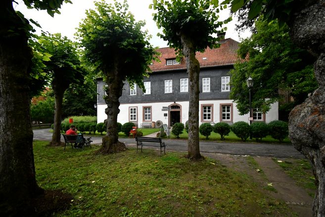 Historisches Gebäude mit Schieferfassade und rotem Ziegeldach, umgeben von Bäumen und Bänken im Vordergrund.