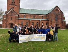 Eine Gruppe von Menschen steht vor einer Kirche und hält ein Banner hoch. Der Banner zeigt Informationen zu einer Veranstaltung in der Innenstadt.