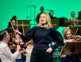 Eine Frau singt leidenschaftlich vor einem Orchester während einer Probe.